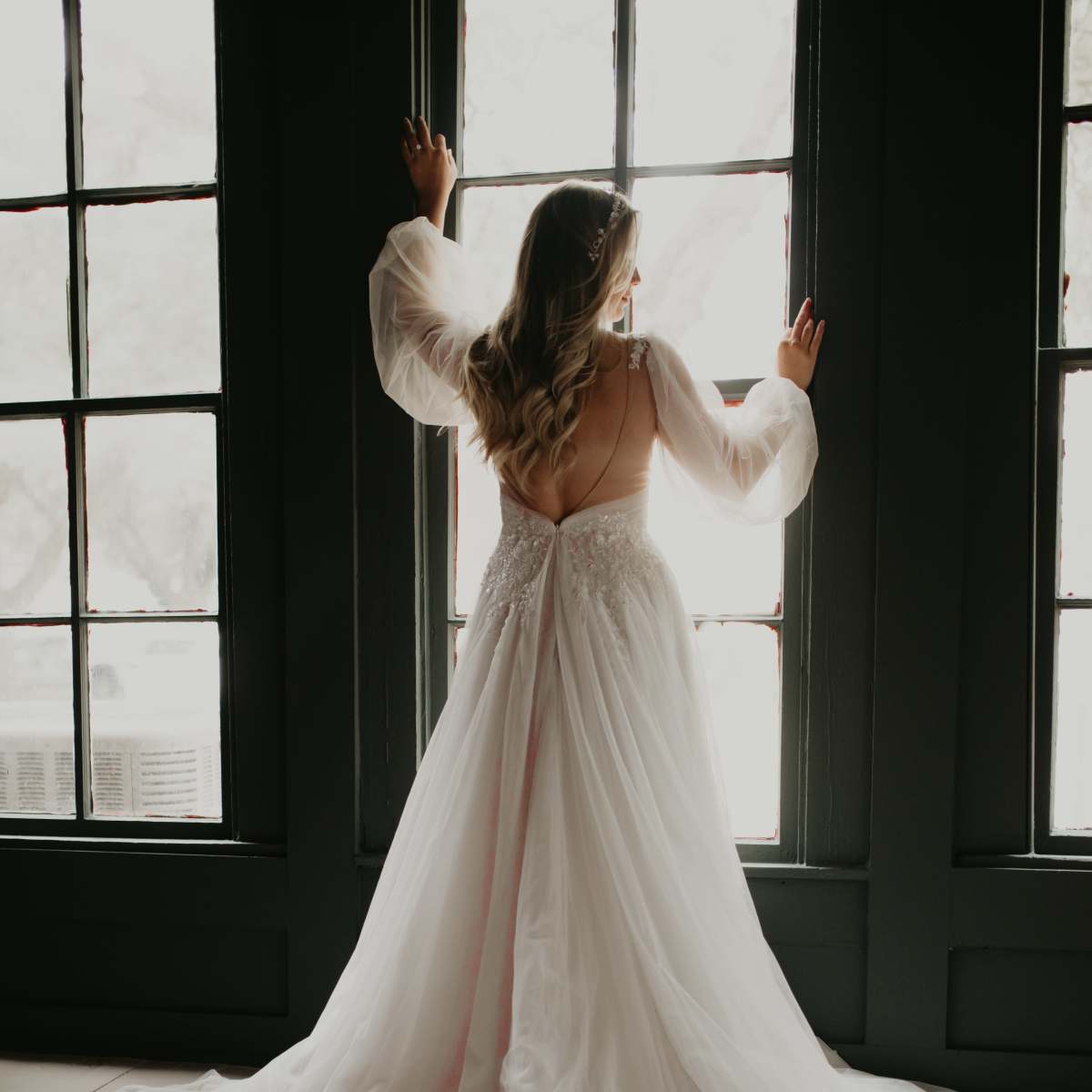 A woman in a flowing white wedding dress stands by a large window, looking outside while holding onto the window frame.