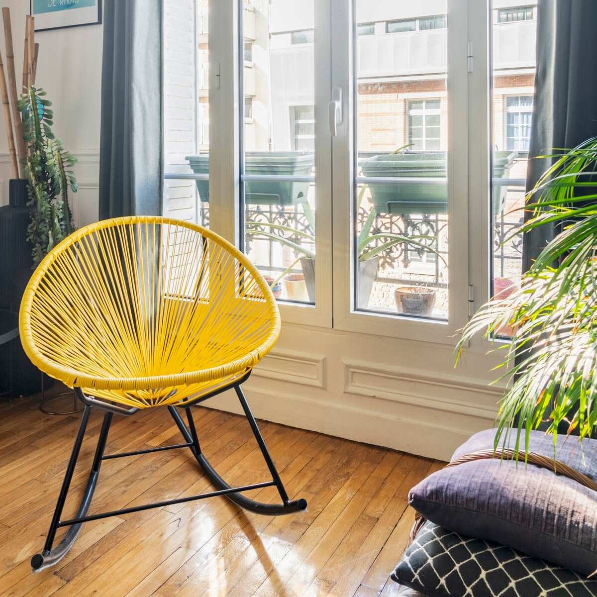 A bright yellow chair is positioned near a large window with curtains, overlooking a balcony filled with greenery.