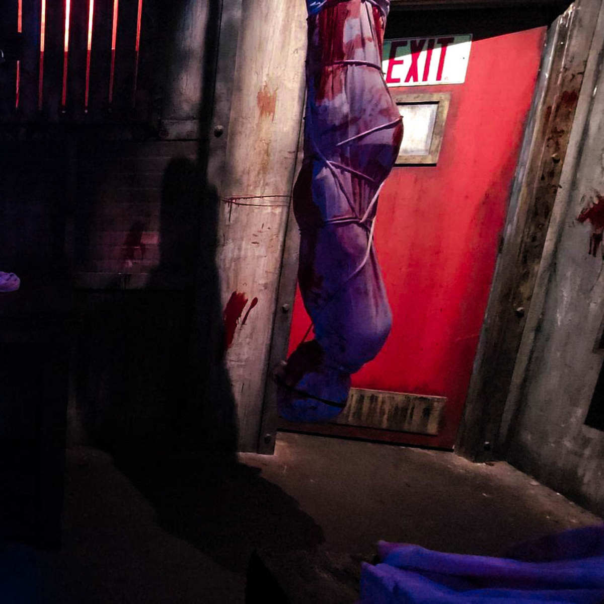 The dimly lit room features a bloodstained wall, a table with a white cloth, and a door marked with an exit sign.