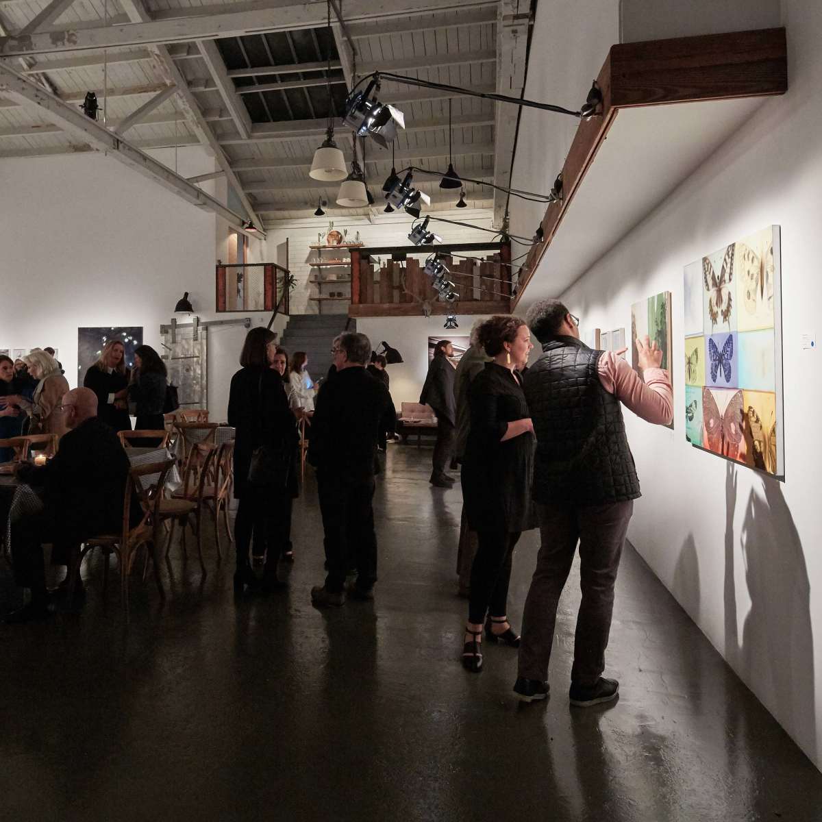 A gallery event is taking place, with several attendees viewing artworks on the wall while others gather around tables in the background.