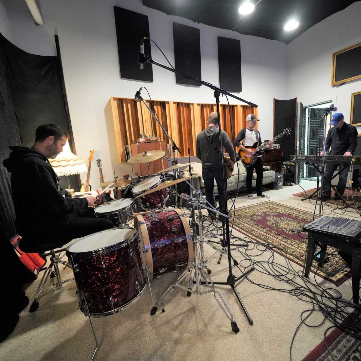 A band consisting of four musicians is rehearsing in a recording studio, with a drummer in the foreground and others playing guitar and keyboards.