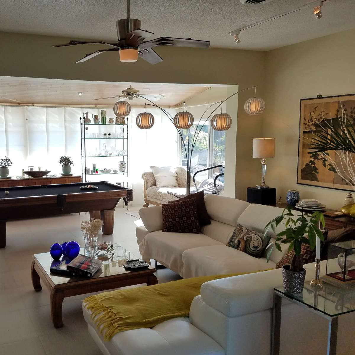 A spacious living area with a pool table, comfortable seating, decorative plants, and large windows allowing natural light to enter.