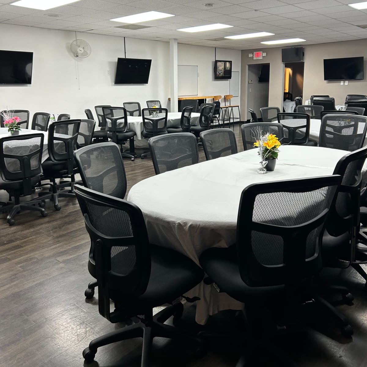 A conference room with multiple circular tables, each covered with a tablecloth, surrounded by ergonomic office chairs and equipped with mounted televisions on the walls.