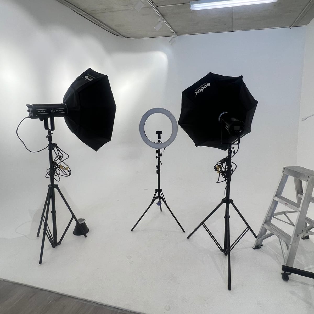 A photography studio setup with two softboxes, a ring light, and a ladder positioned against a plain white backdrop.