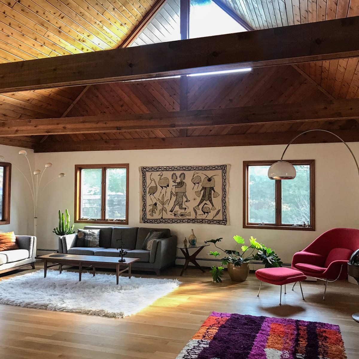 A spacious living room features a high wooden ceiling, large windows, a modern sofa set, and decorative plants.