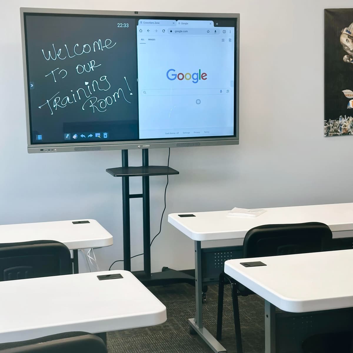 A digital display in a classroom shows a welcome message alongside a Google search page, with empty desks arranged in front.