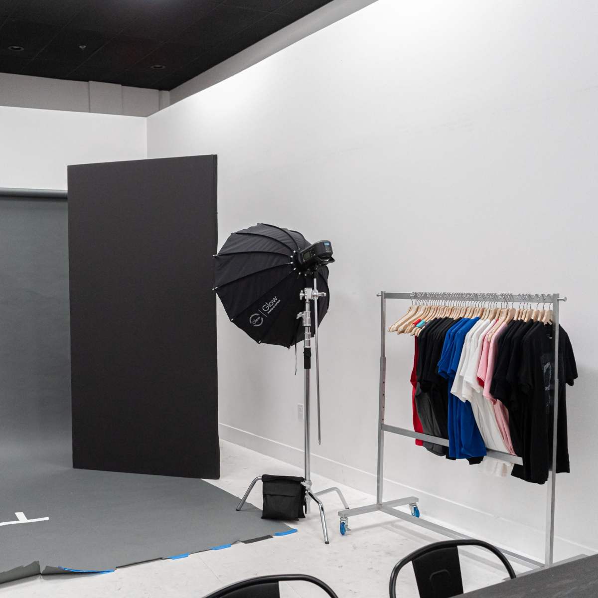 A photography studio setup with two lighting umbrellas, a backdrop, and a rack of assorted t-shirts in various colors.