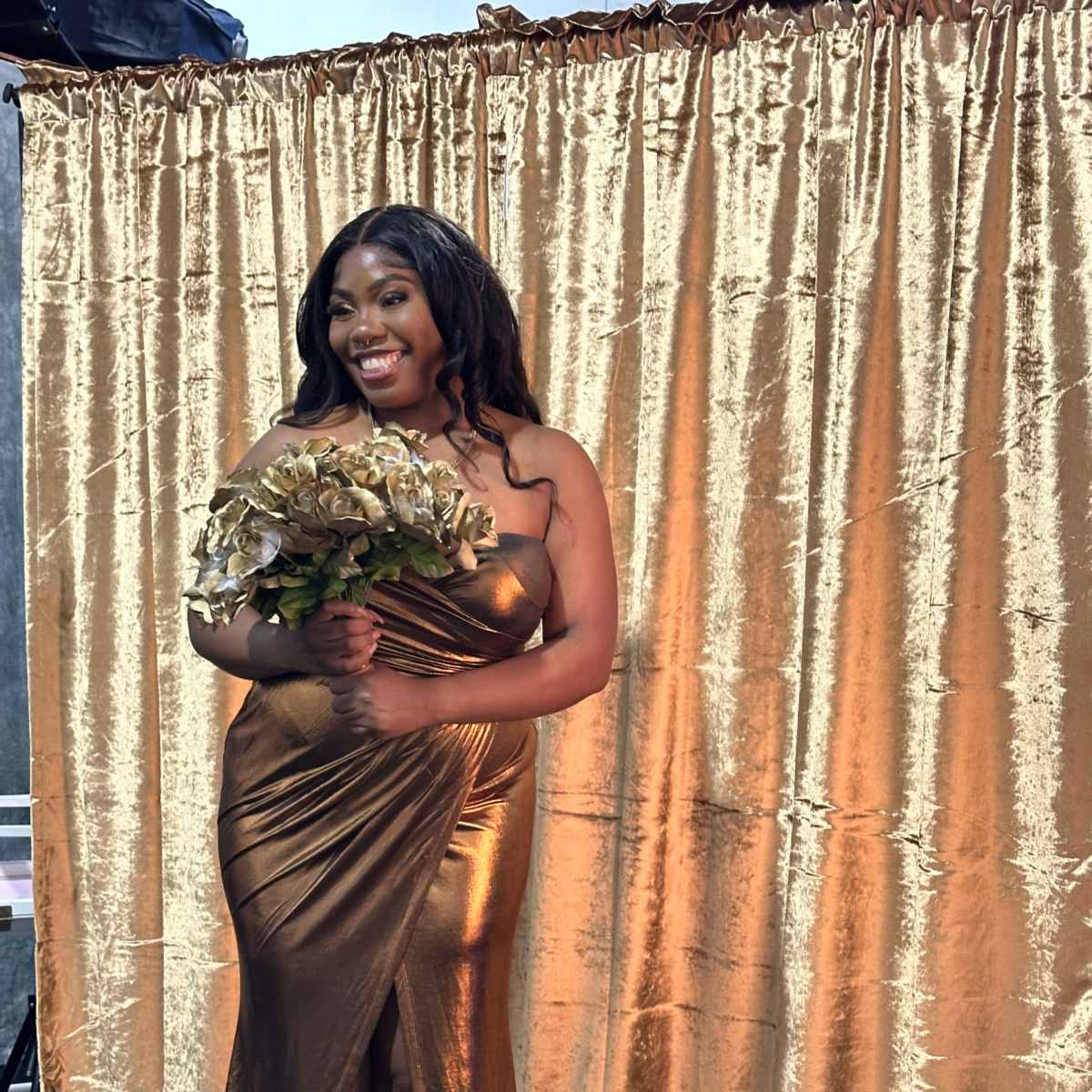 A woman in a gold dress holds a bouquet in front of a gold curtain backdrop.