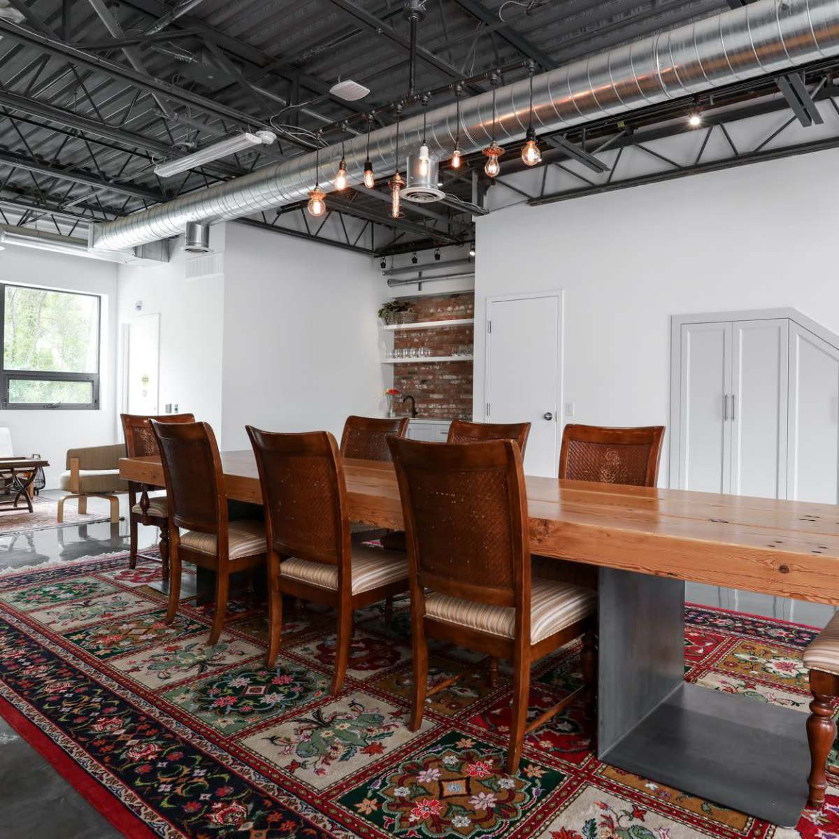 A long wooden dining table is surrounded by several chairs on a patterned rug in a brightly lit room with exposed pipes and a staircase.