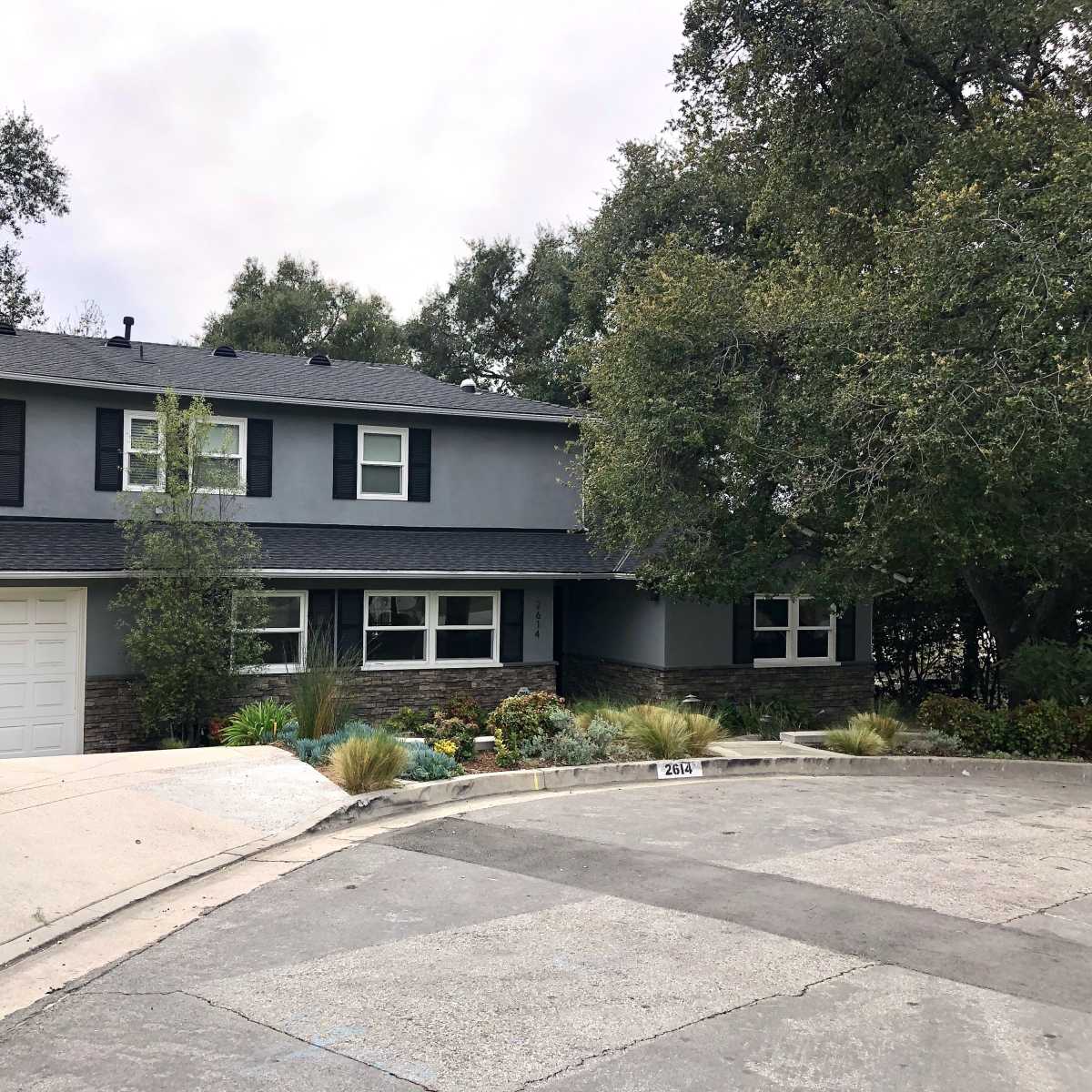 A two-story gray house with a stone facade sits at the end of a circular driveway surrounded by trees and low shrubs.