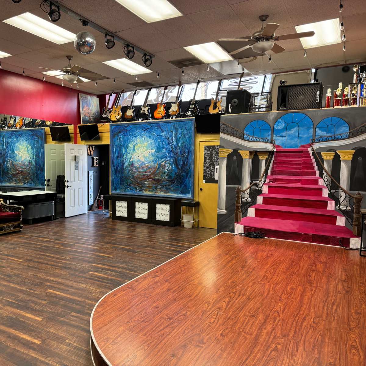 The interior of a dance studio features a polished wooden floor, red walls with artwork, large mirrors, a staircase mural, and musical instruments on display.