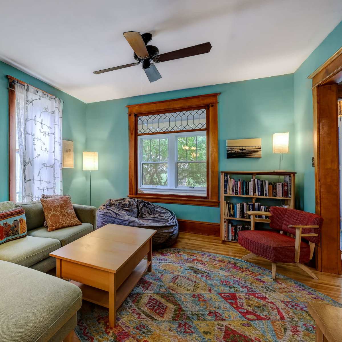 A cozy living room featuring a green wall, a sectional sofa with decorative pillows, a wooden coffee table, and a window with patterned curtains, accompanied by bookshelves and a bean bag chair.