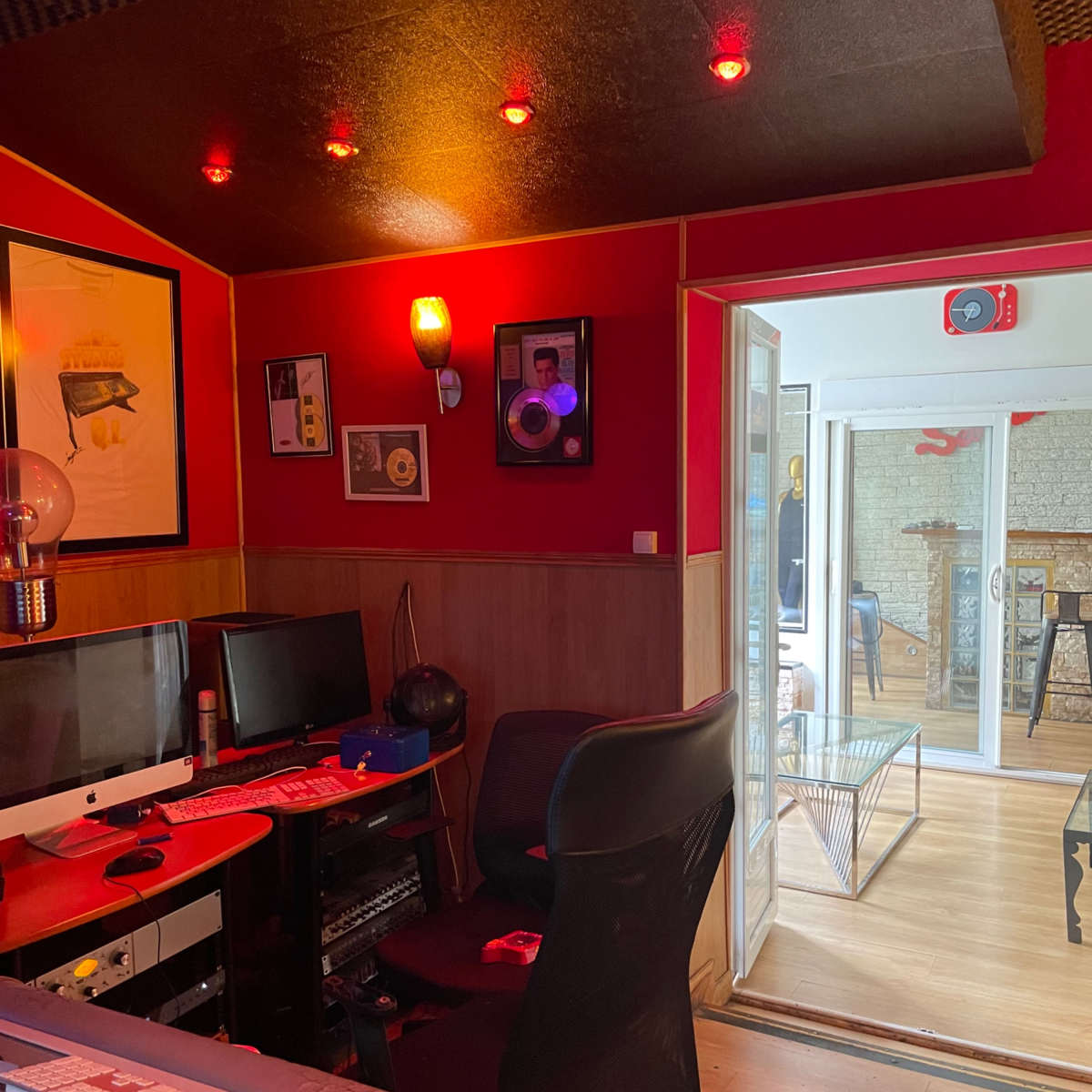A dimly lit recording studio with red walls, a mixing console in the foreground, and a glass door leading to a bright, multicolored space.