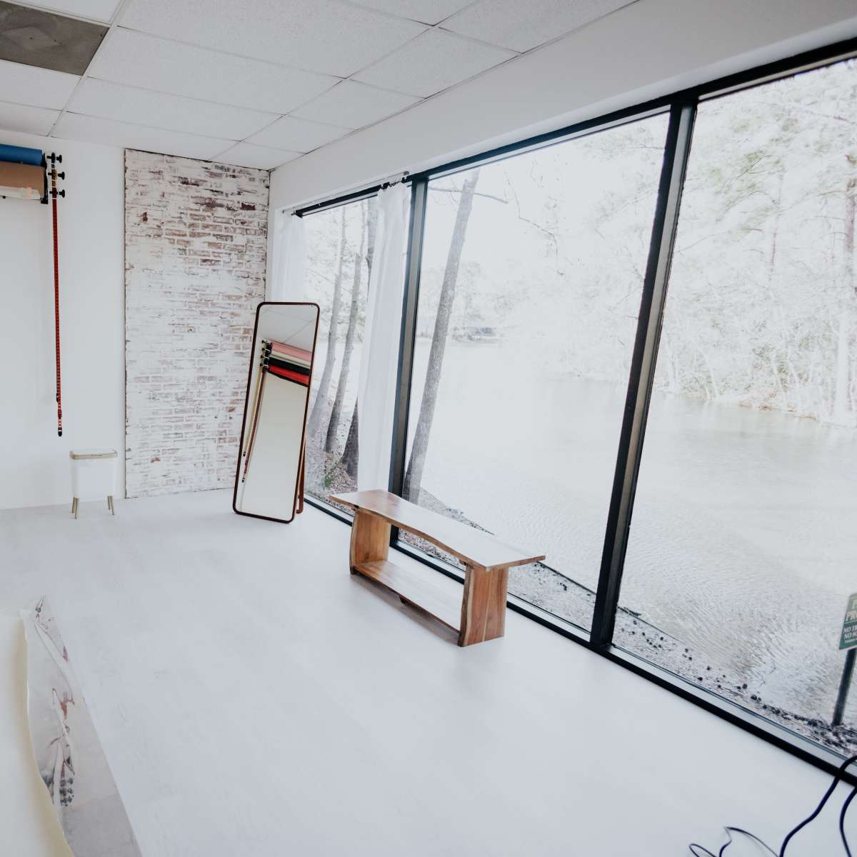 A brightly lit, minimalistic room features a large window overlooking a body of water, with a wooden bench and a standing mirror inside.