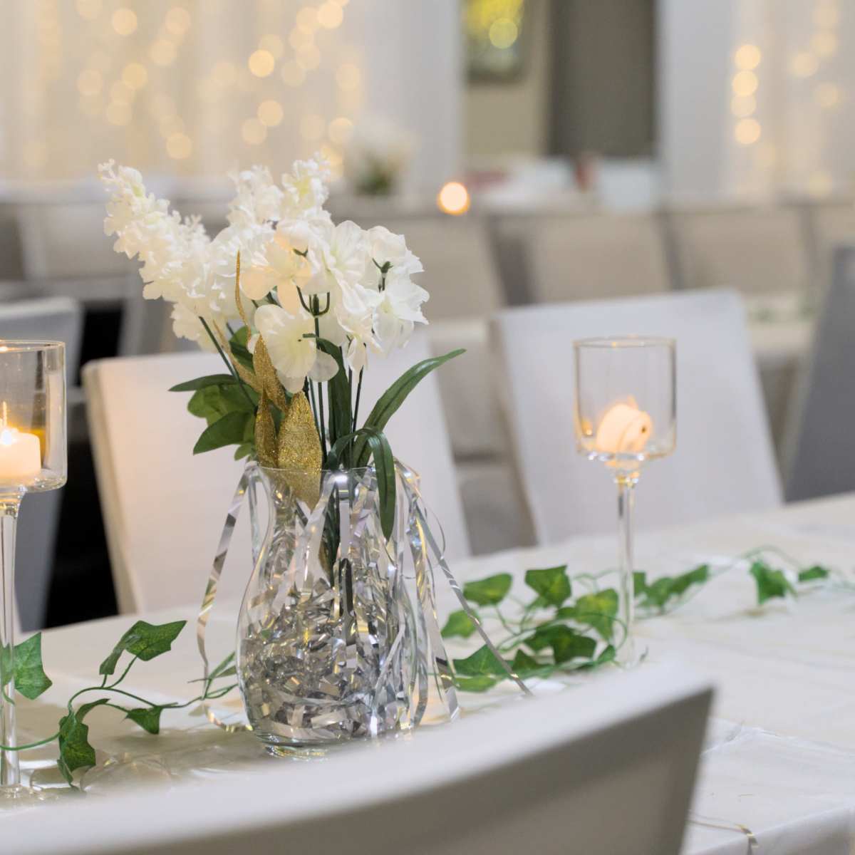 A table is set with a floral centerpiece, candles, and decorative greenery in a softly lit dining area.