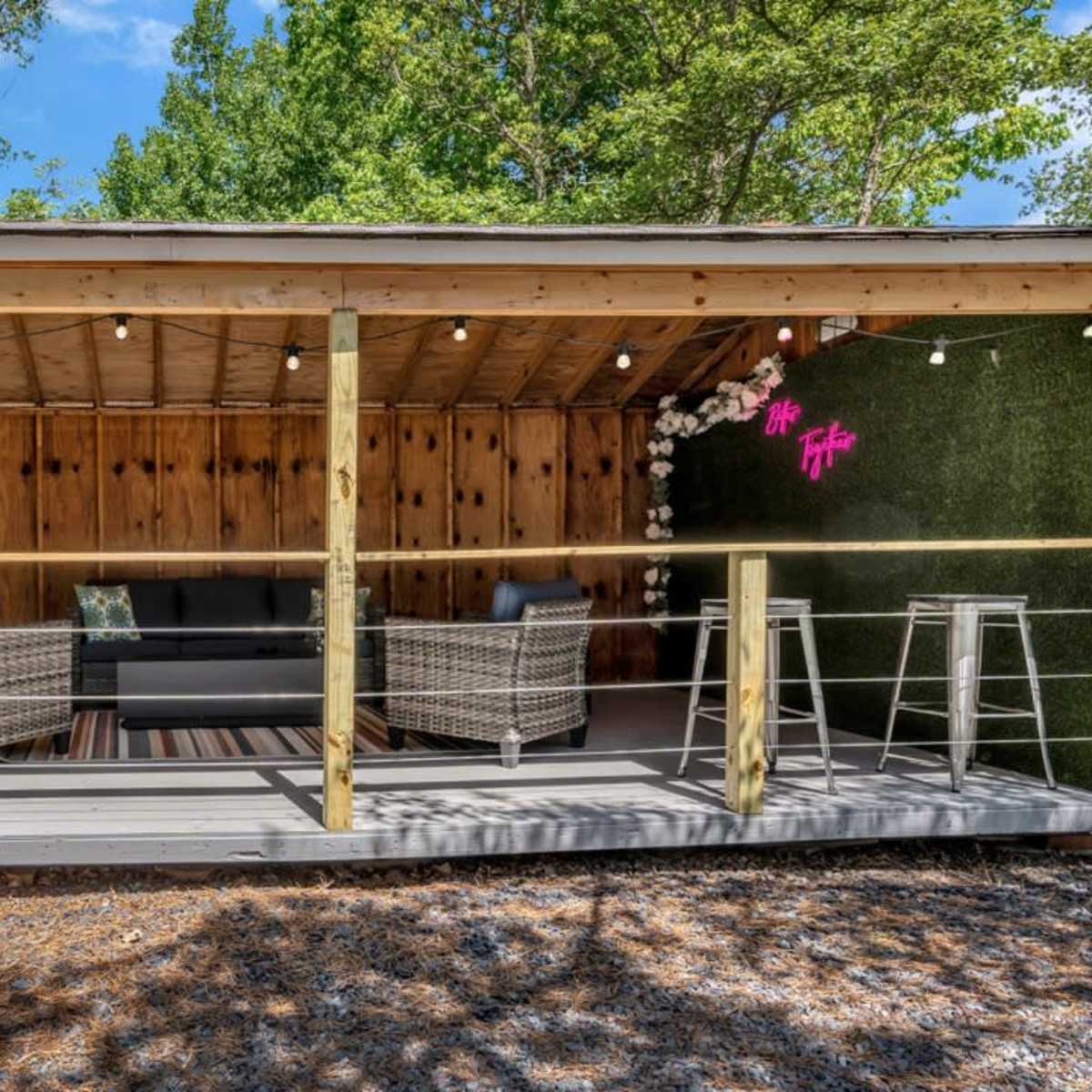 A modern outdoor lounge area with two wicker chairs, a sofa, and decorative plants under string lights, set within a wooden structure.