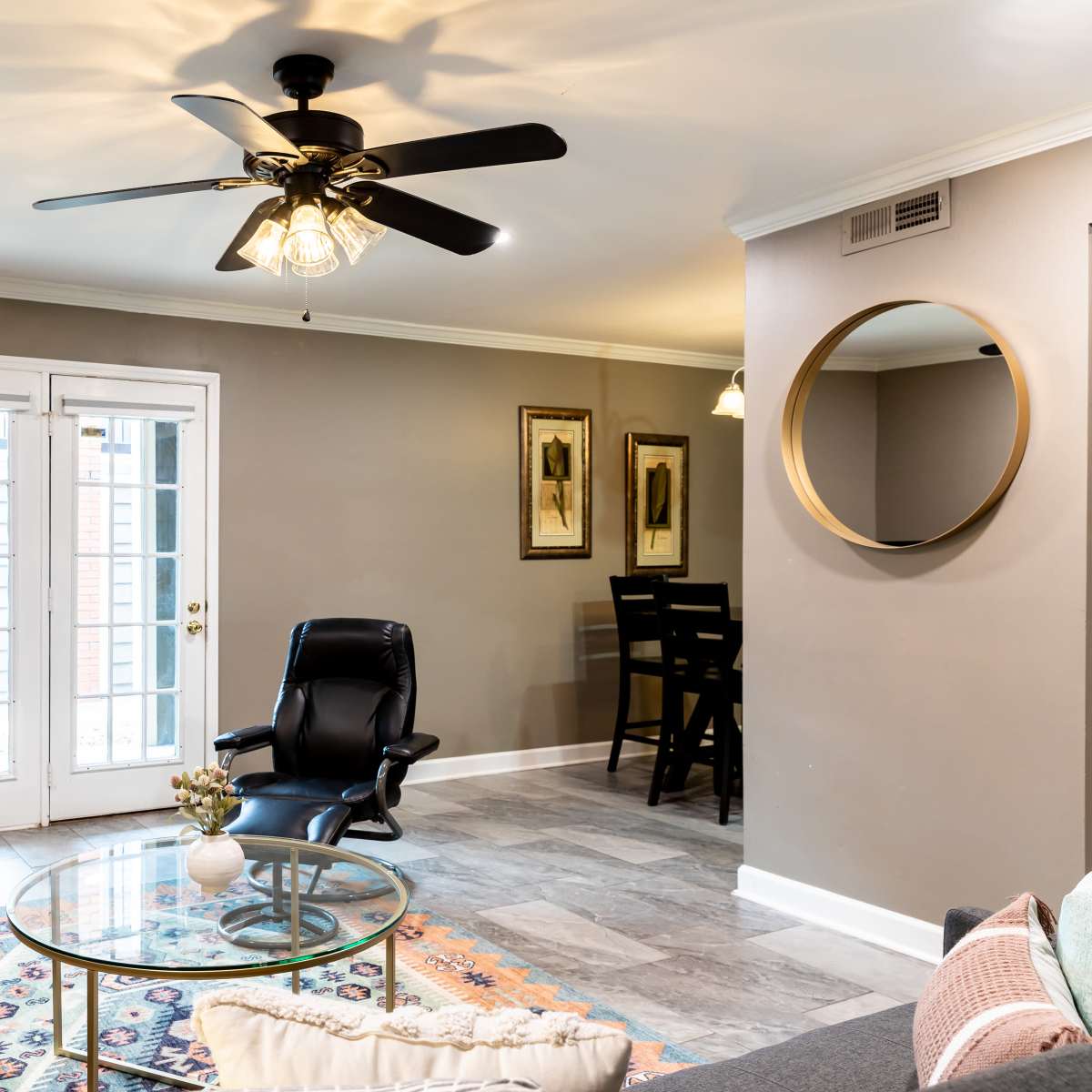 A modern living room with a ceiling fan, a black leather recliner, a glass coffee table, and a TV, with sliding glass doors leading to an outdoor area.