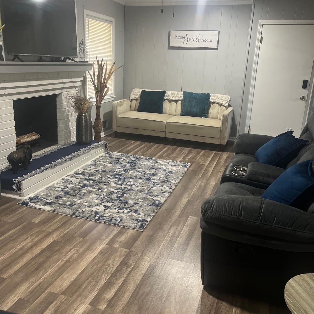The living room features a light-colored sofa with blue cushions, a dark recliner, a patterned rug, and a television mounted above a white brick fireplace.