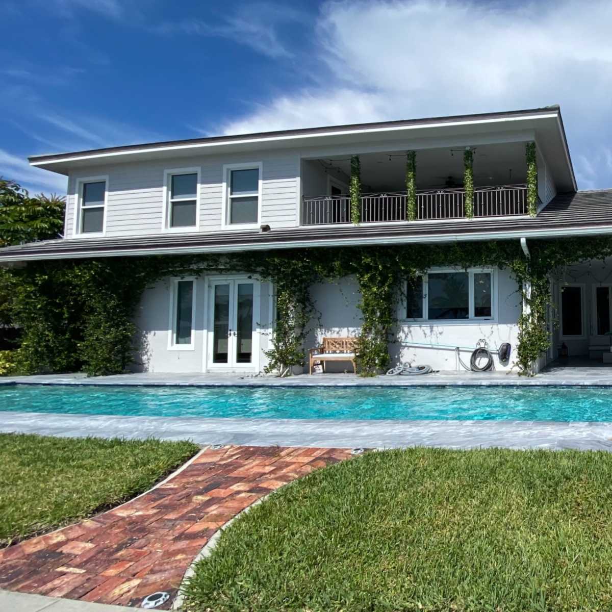 A two-story house with a pool in the foreground is surrounded by green grass and climbing vines on the walls.
