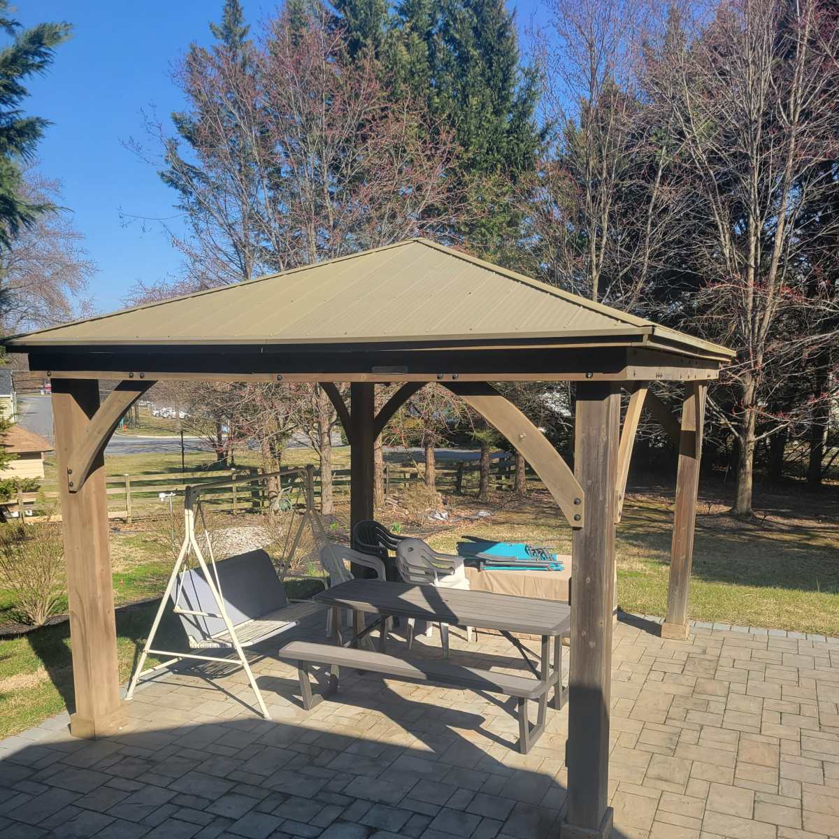 A wooden gazebo with a metal roof stands on a stone patio surrounded by trees and grass.