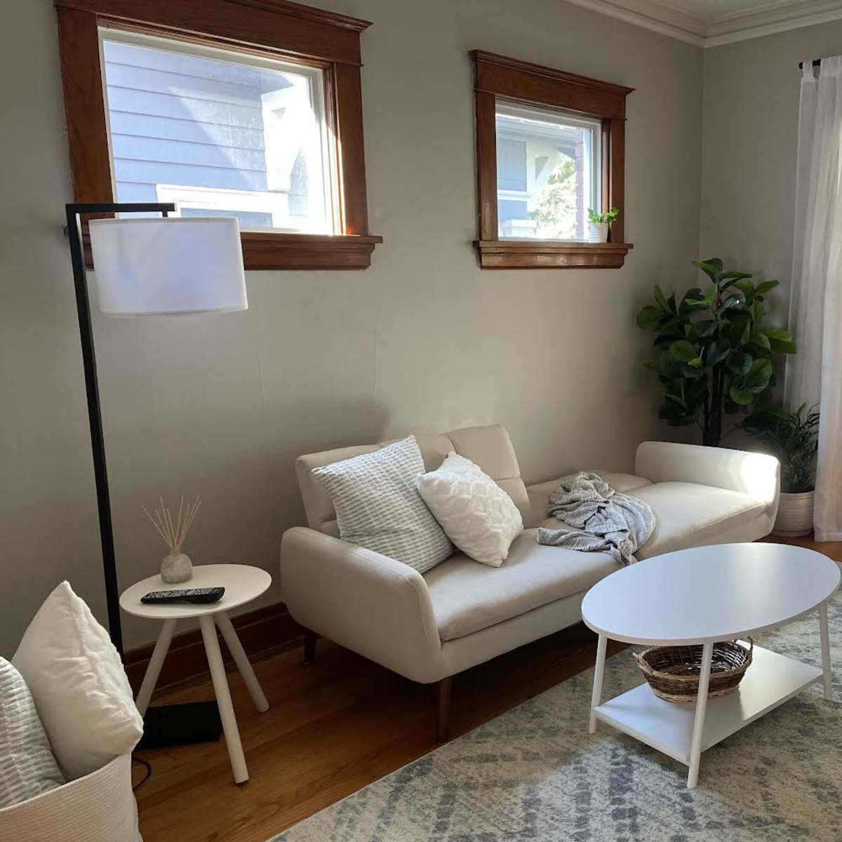 A cozy living room with a white sofa, a small round table, and large windows allowing natural light to enter.