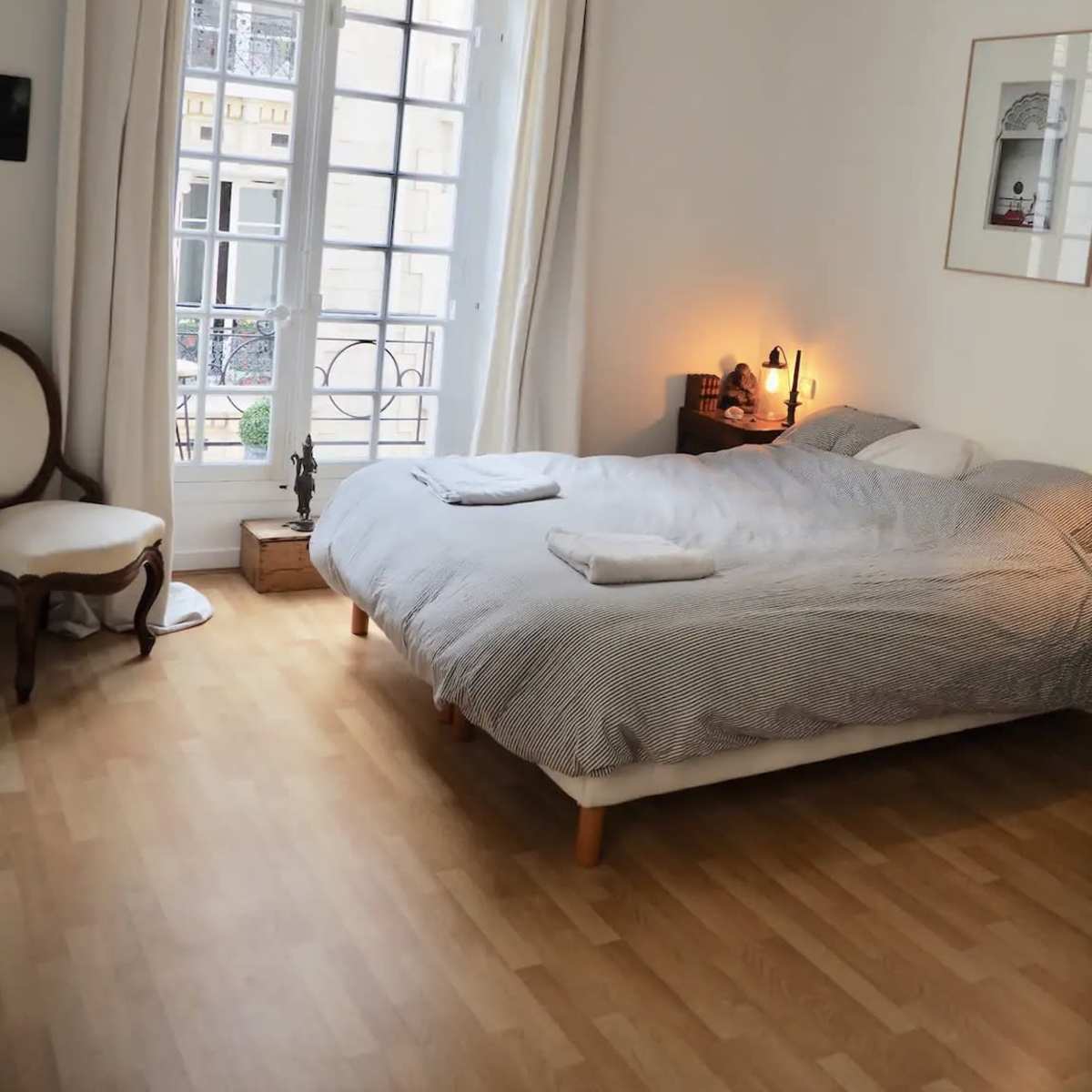 A neatly arranged bedroom featuring a large bed with gray bedding, a small nightstand with a lamp, and a window adorned with light curtains.