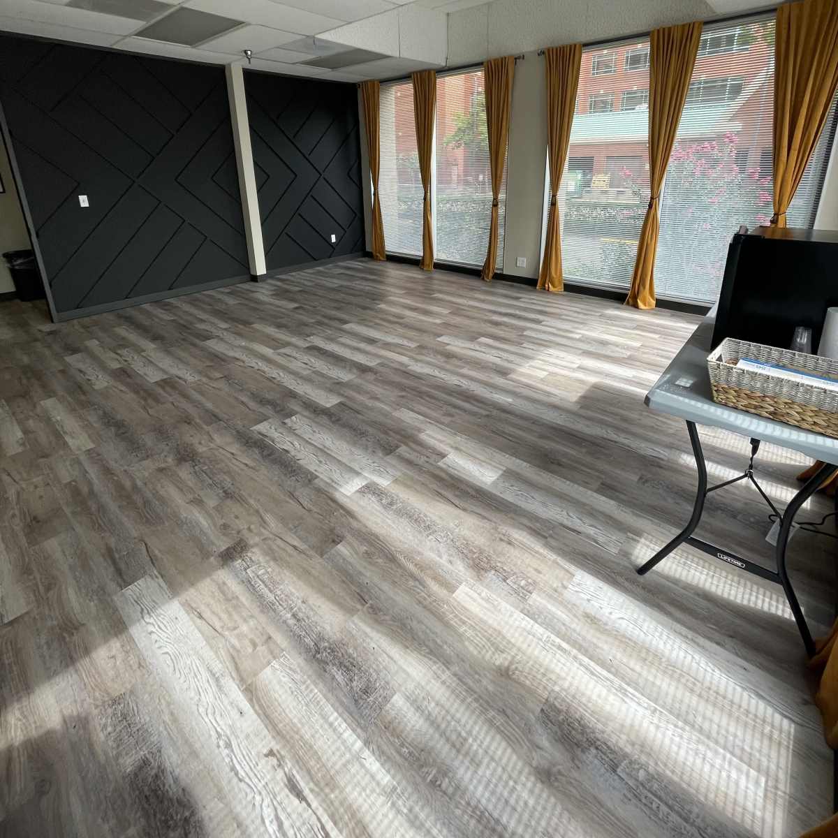 A spacious interior room with large windows and wooden flooring, featuring orange curtains along the windows.