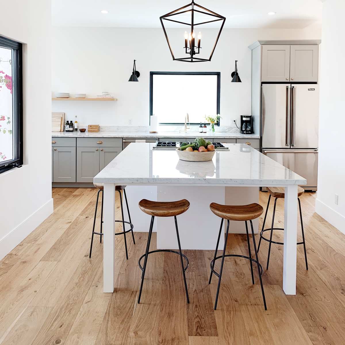 A modern kitchen features a large island with three stools, gray cabinetry, stainless steel appliances, and ample natural light from a window.