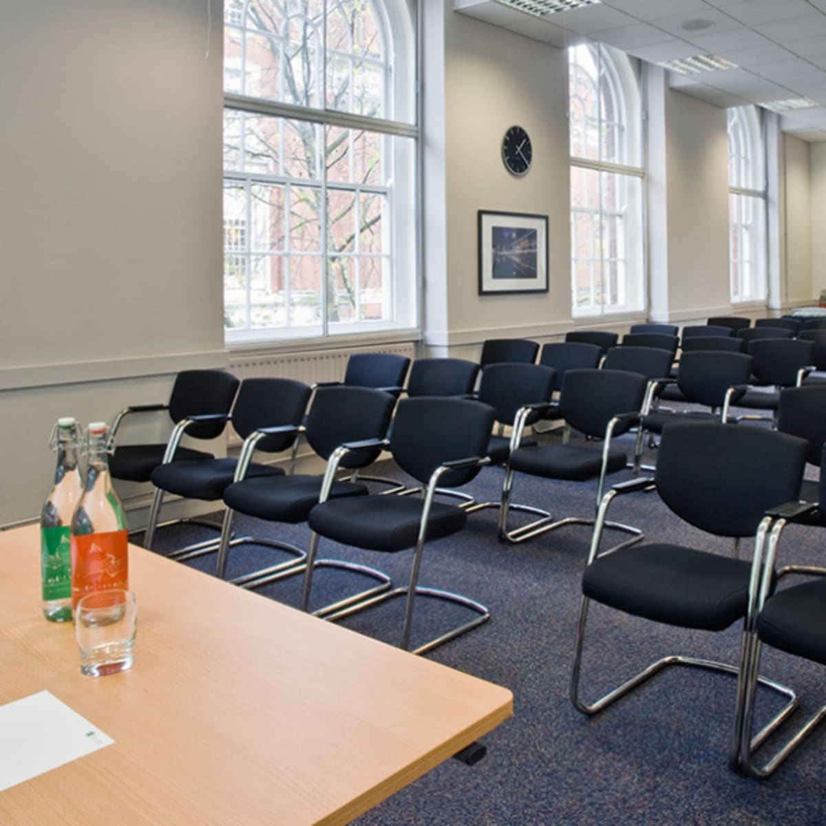 A meeting room with several rows of empty black chairs facing a small table with two bottles of water and glasses.
