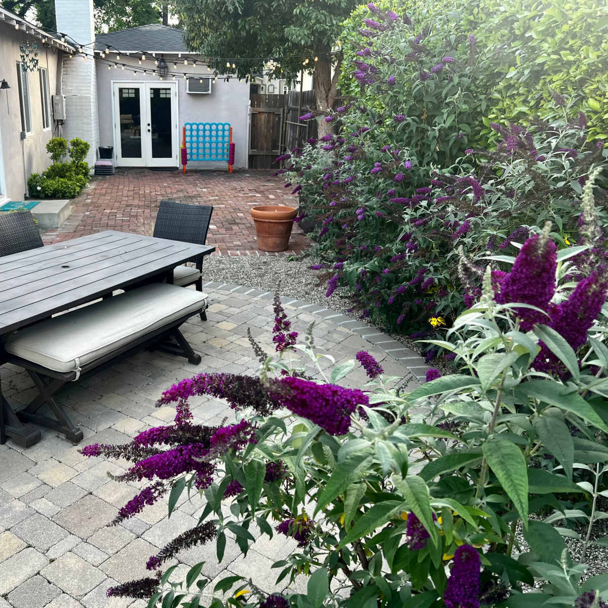 A wooden dining table with chairs is positioned on a stone patio surrounded by flowering plants and a brick pathway leading to a nearby building.