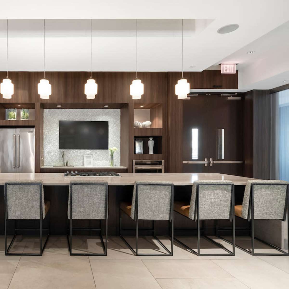A modern kitchen features a long island with eight high-backed stools and a wall-mounted television above a sleek backsplash.