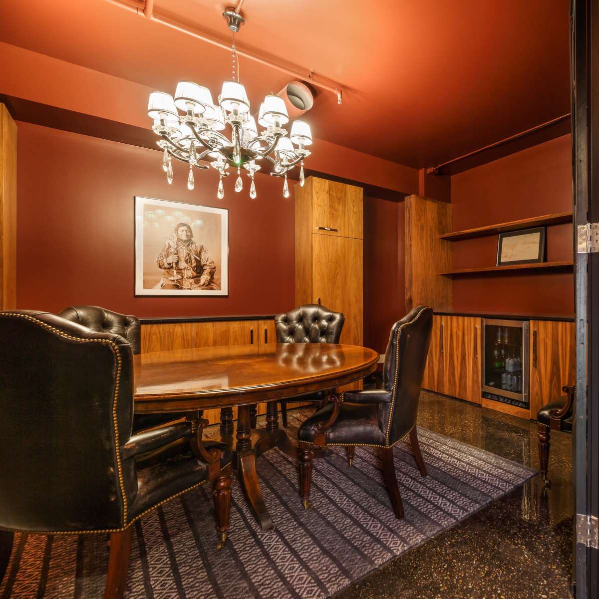 A conference room features a round wooden table surrounded by high-backed chairs, with a chandelier overhead and dark red walls.