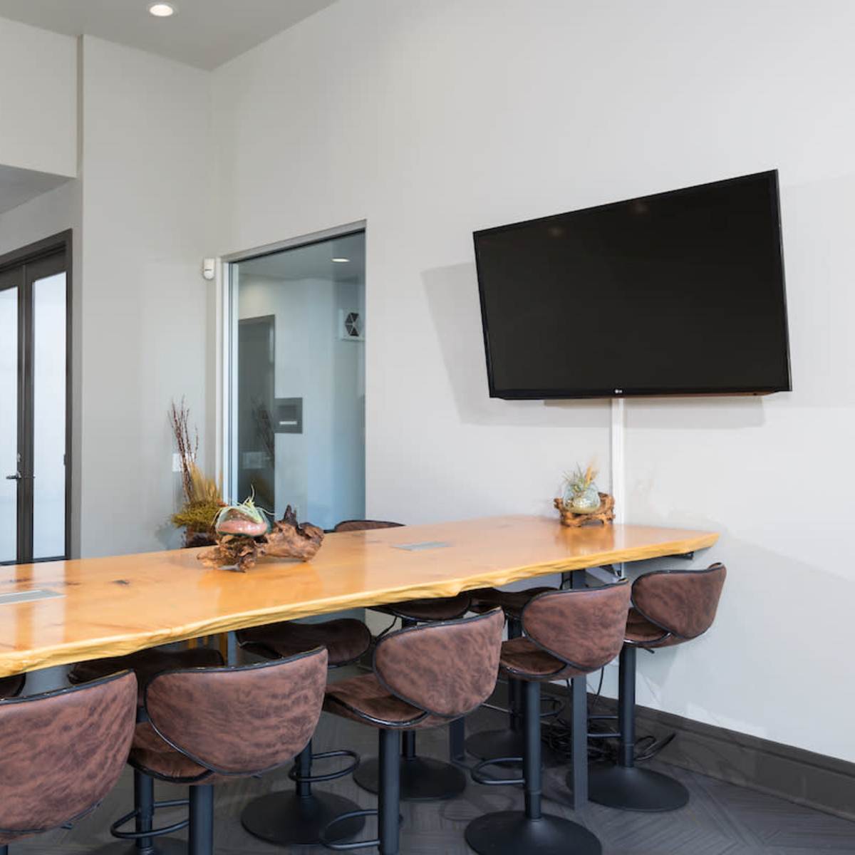 A modern conference room features a long wooden table with stools, a wall-mounted television, and decorative elements on shelves.