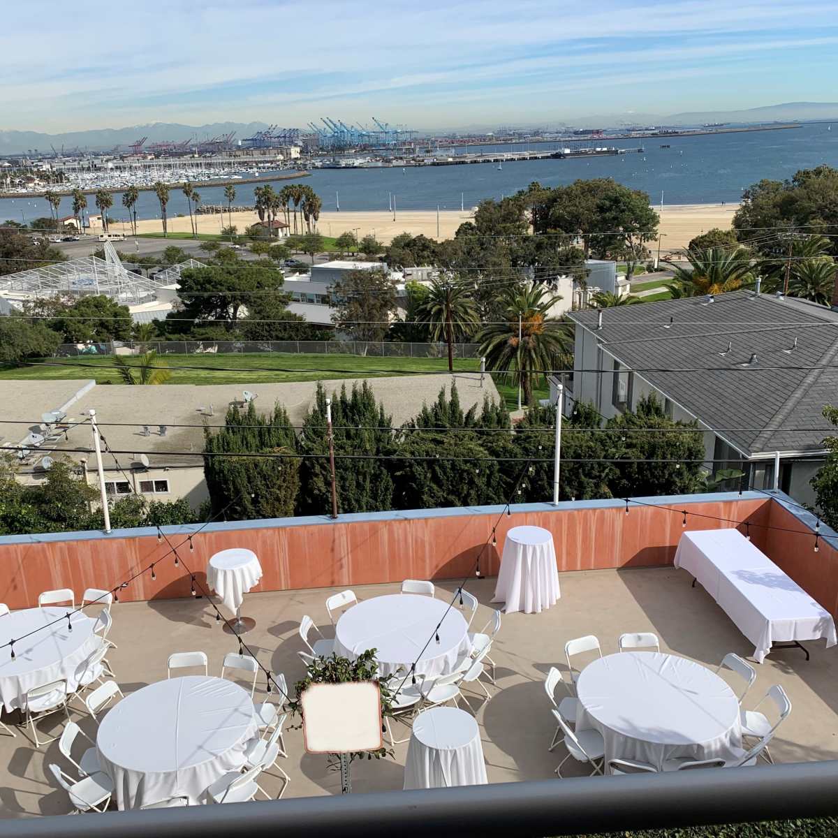 A terrace with several round tables and chairs set up for an event, overlooking a coastal view with palm trees and distant shipping docks.