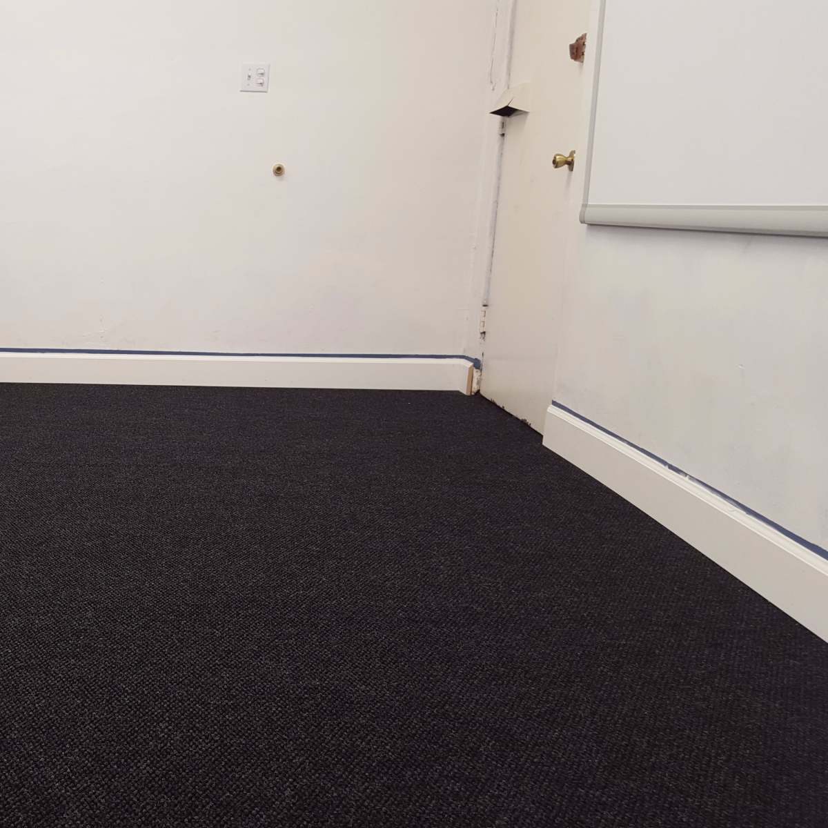 An empty room with dark carpet, a black piano in the corner, and a door on the right.