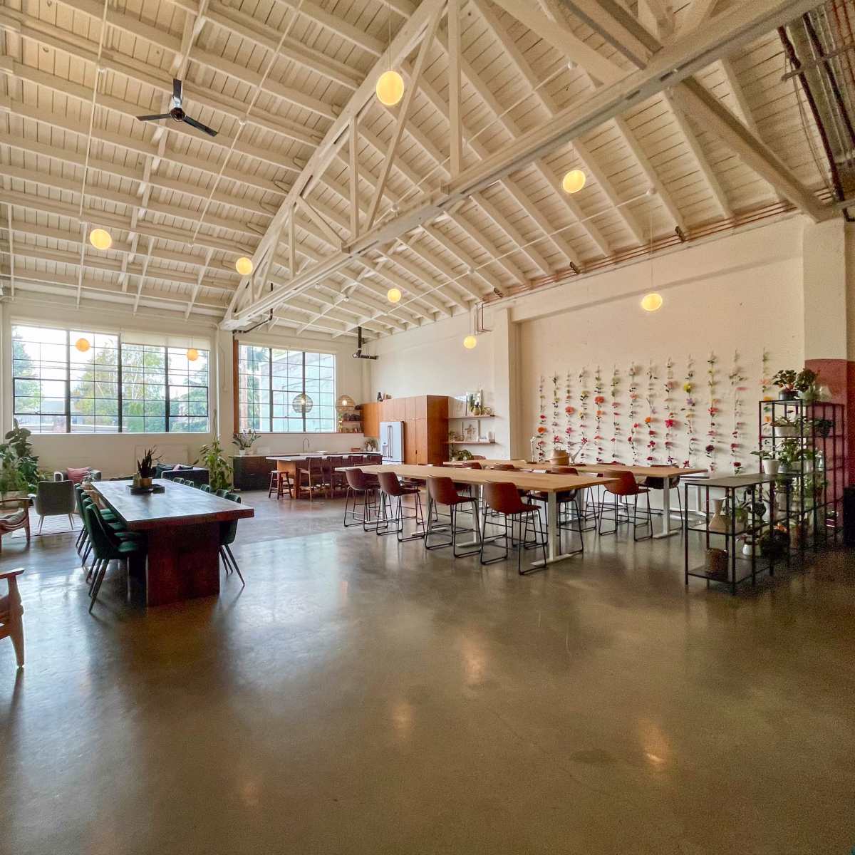 A spacious, bright interior of a dining or workspace with high ceilings, large windows, and various seating arrangements.