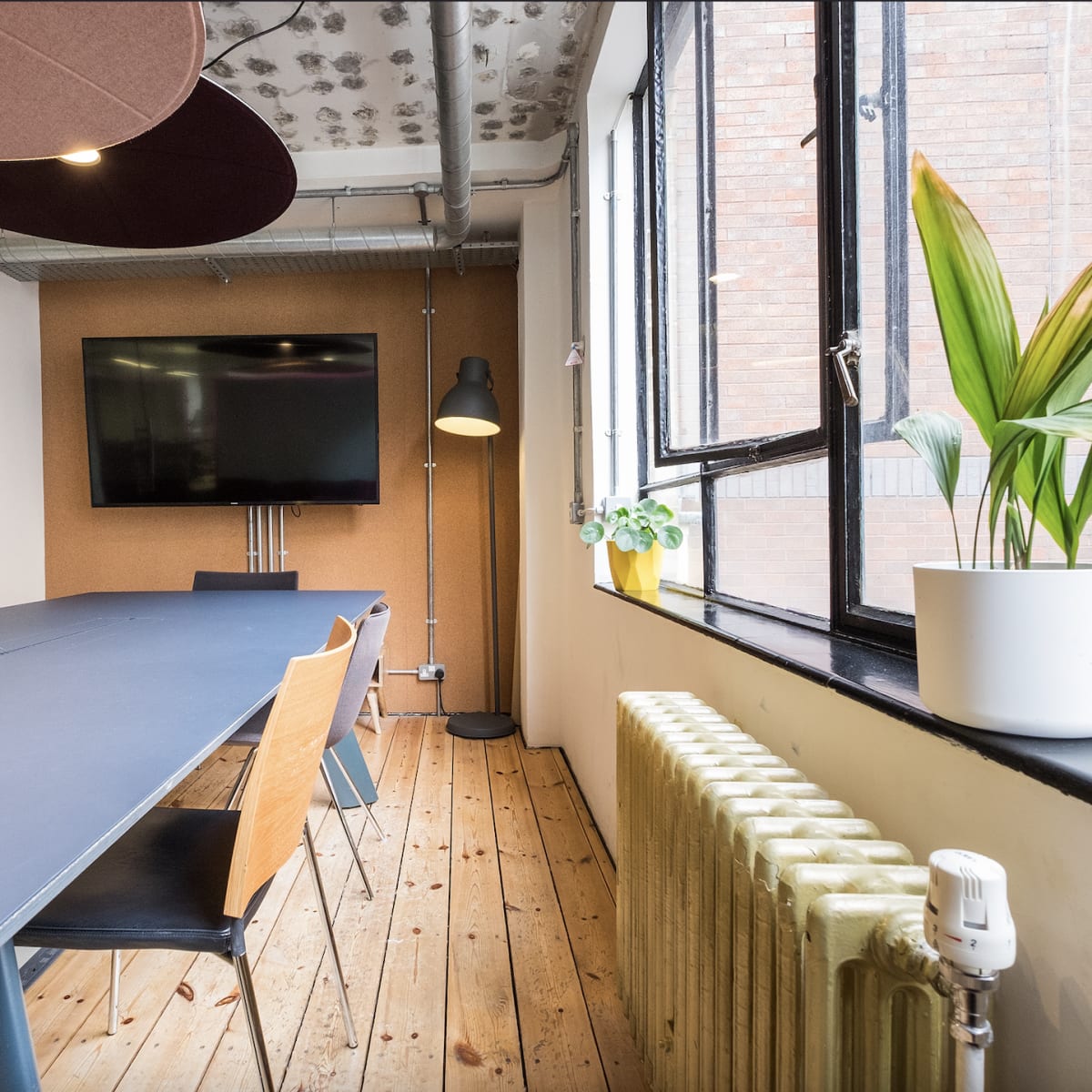 A modern conference room features a long table, several wooden chairs, a wall-mounted television, and a window with a potted plant beside a radiator.