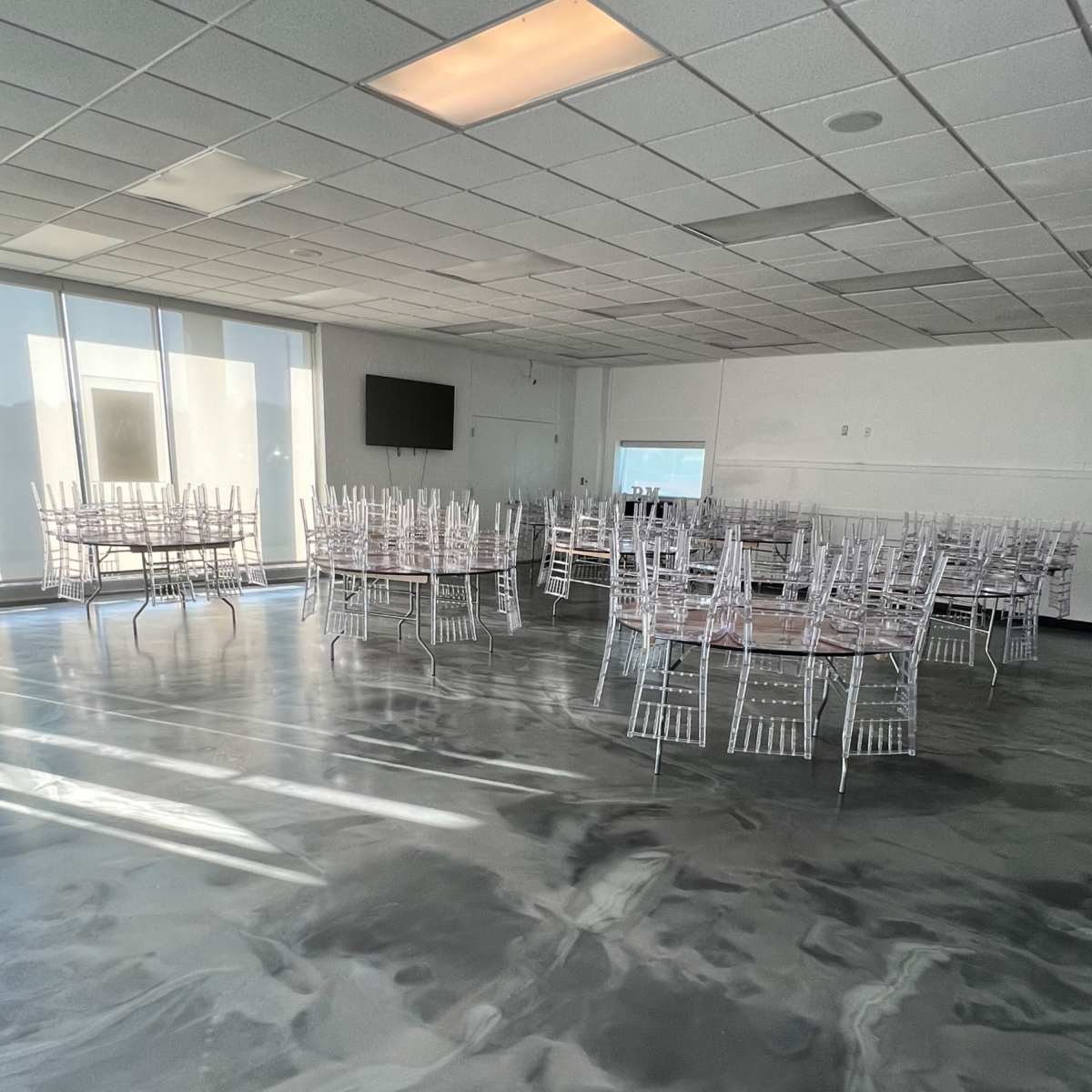 A spacious room with several round tables set up, each surrounded by transparent chiavari chairs, on a polished floor.