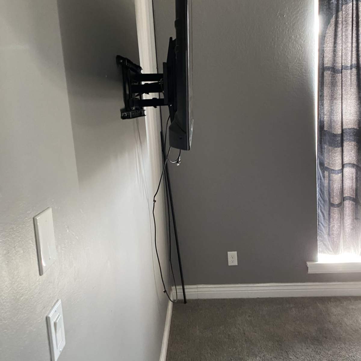 A flat-screen television is mounted on a gray wall, with a curtain partially covering a window nearby.