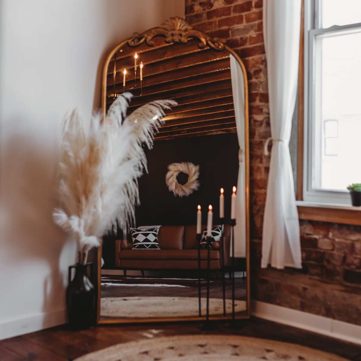 A tall, ornate mirror reflects a cozy seating area with a plant and candles in a warmly lit room featuring exposed brick walls.