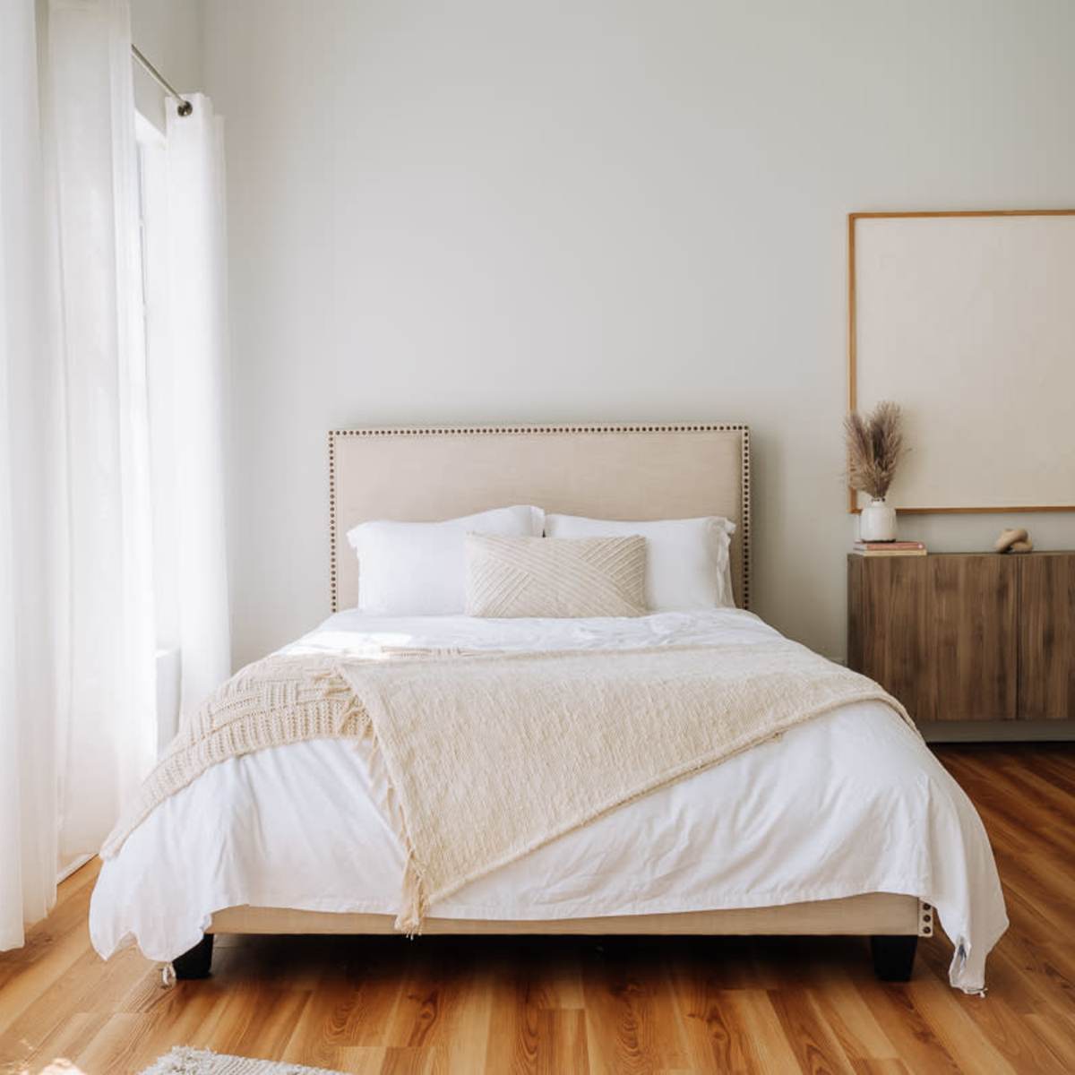A bright bedroom featuring a bed with white bedding, a light-colored headboard, and a wooden dresser beside a large window with sheer curtains.