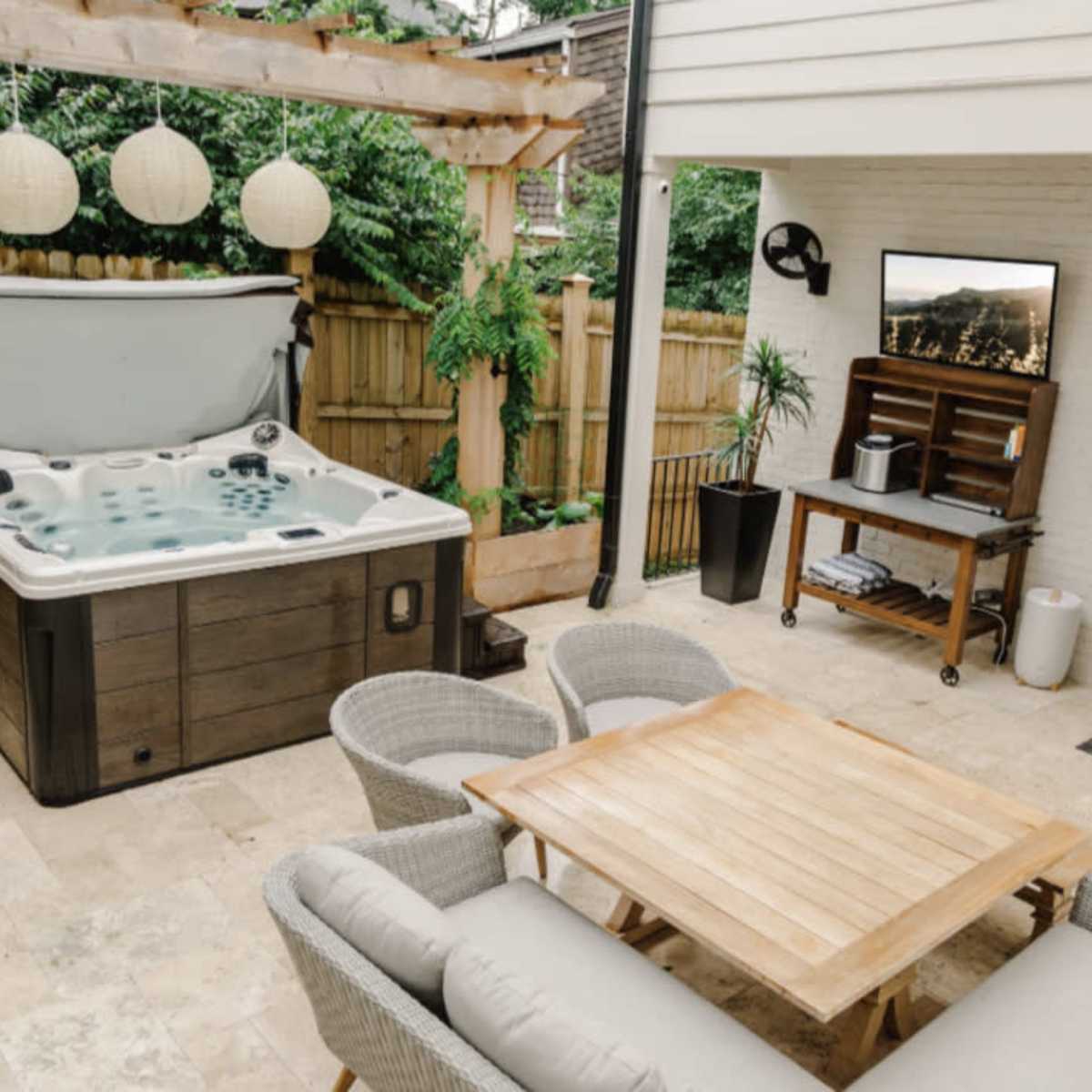 An outdoor patio area featuring a hot tub, a wooden table with chairs, and a mounted television, surrounded by greenery and a wooden fence.
