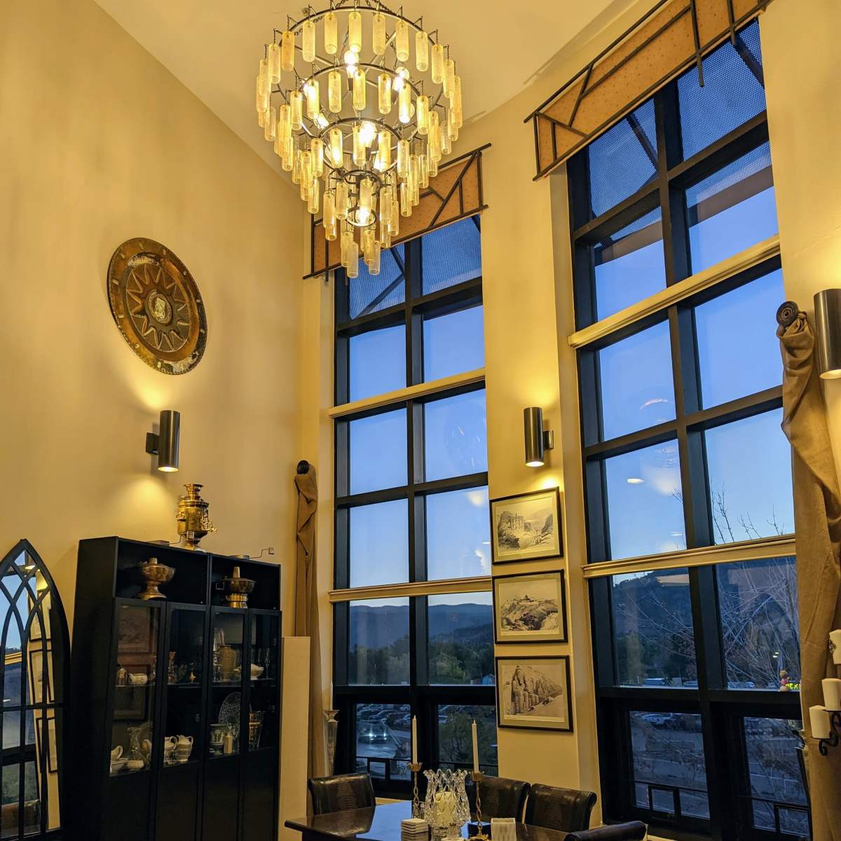 A tall room features large windows, a chandelier, a dining table with chairs, and a display cabinet against the wall.