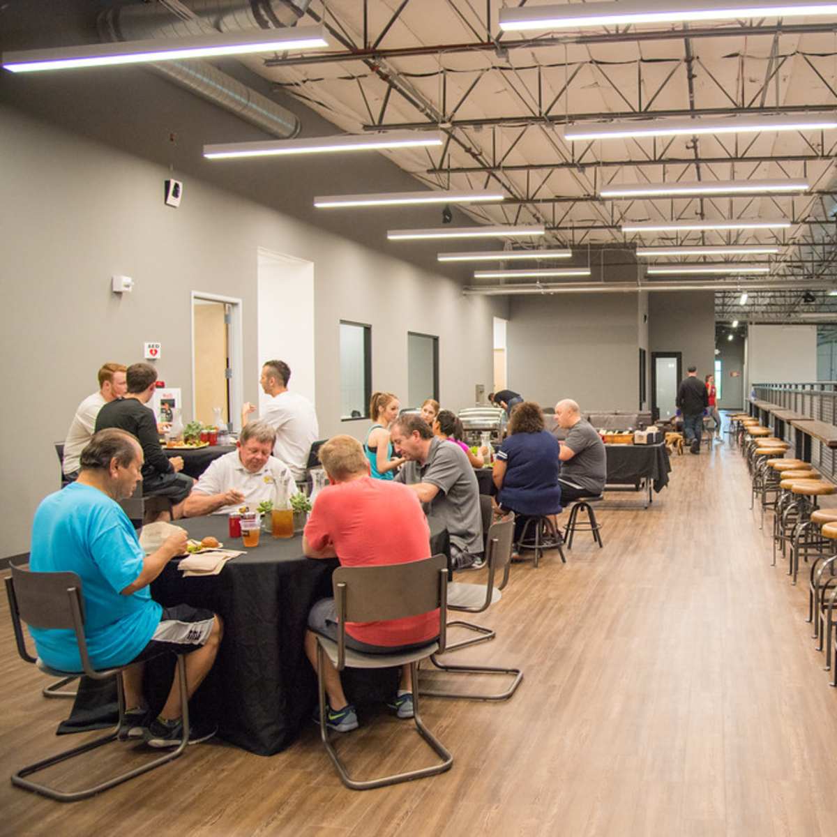 A group of people sits at tables and high-top seating in a spacious indoor venue, sharing a meal and conversation.