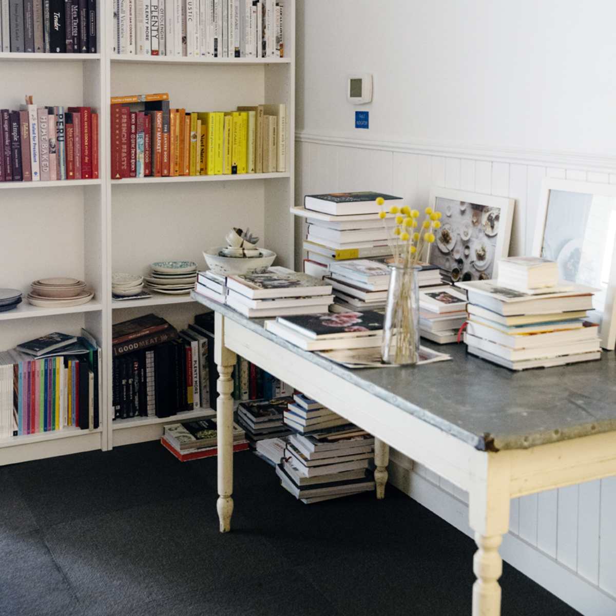 A white-walled room features a bookshelf filled with neatly arranged books, a table stacked with more books, and a vase of yellow flowers.