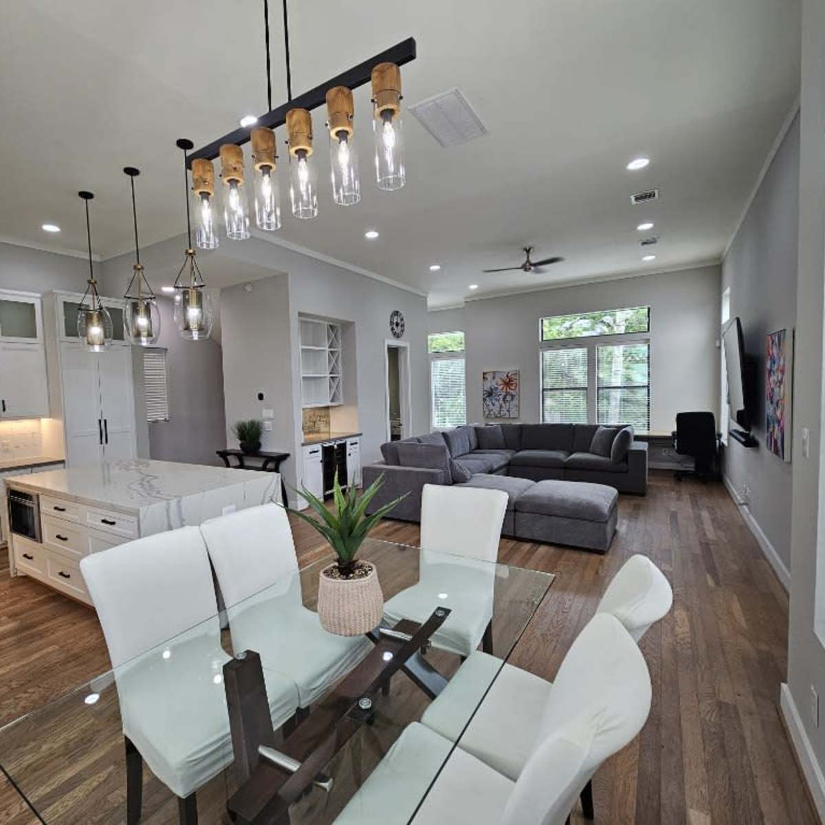 A modern open-concept living space featuring a dining area with a glass table, white chairs, a kitchen with stainless steel appliances, and a living area with a gray sectional sofa.