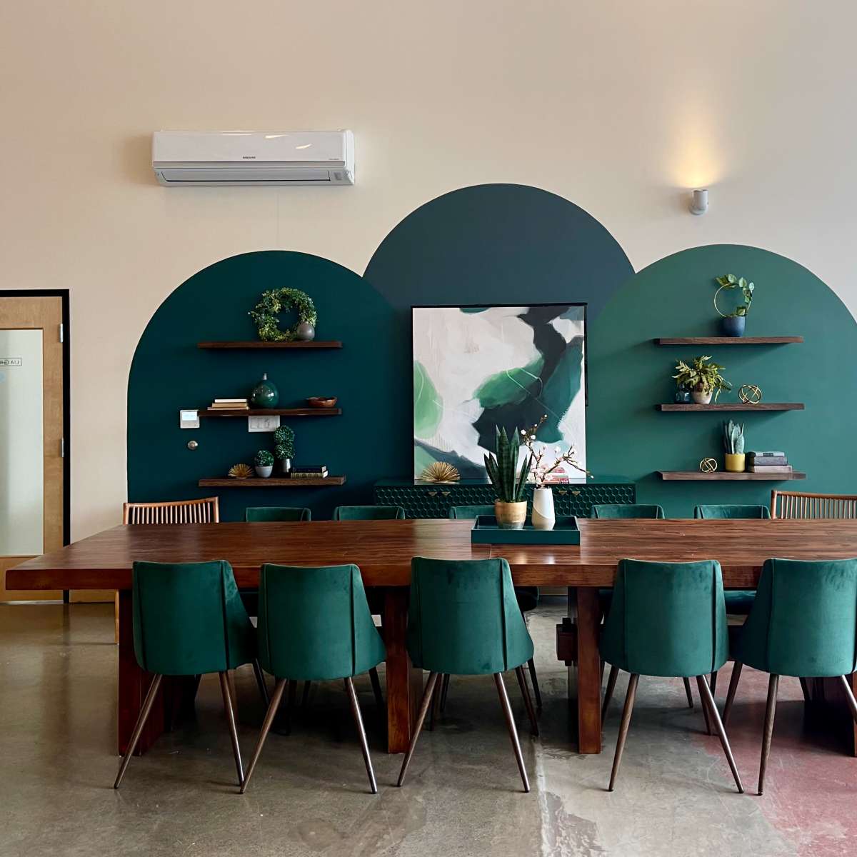 A modern dining area featuring a large wooden table surrounded by green chairs, with a shaded green wall and shelves displaying plants and artwork in the background.