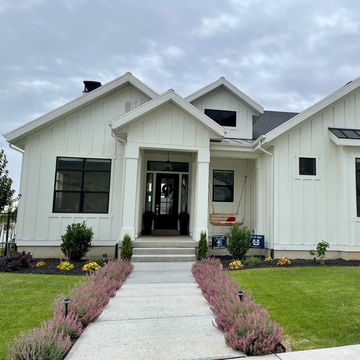 A modern white farmhouse with large windows and a landscaped front yard featuring a pathway and colorful flower beds.
