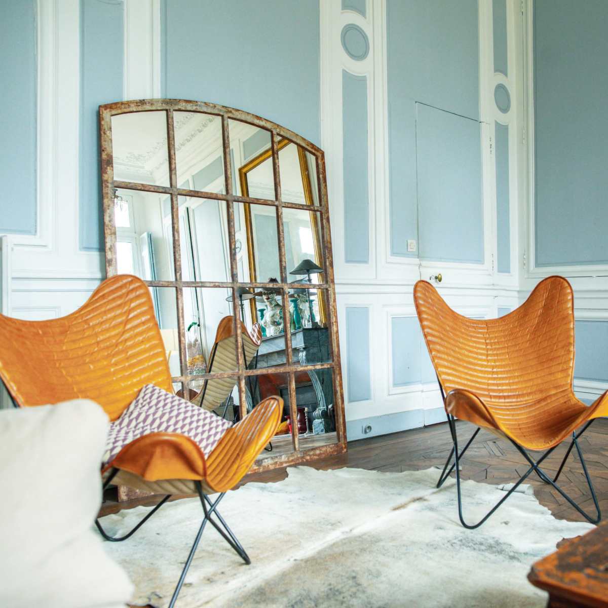 The scene shows a stylish living room with two butterfly chairs, a large mirror, and a decorative rug on the floor.