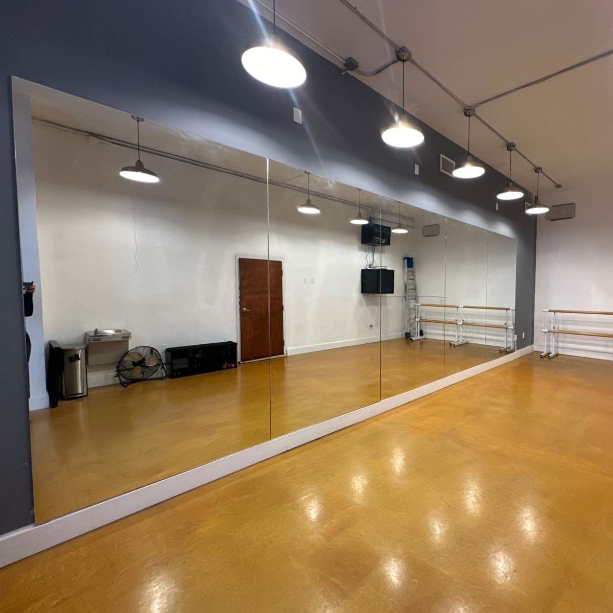 A dance studio featuring a mirrored wall, wooden floors, and overhead lighting.